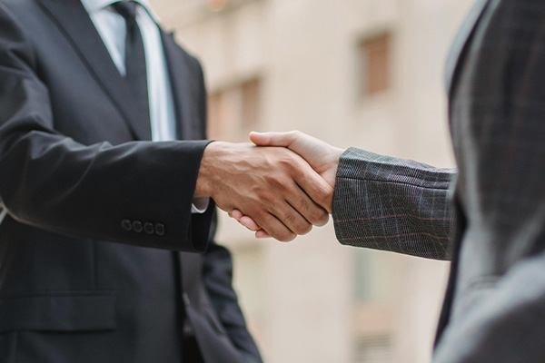 Close-up of two people in suits shaking hands in front of an open laptop on a desk.